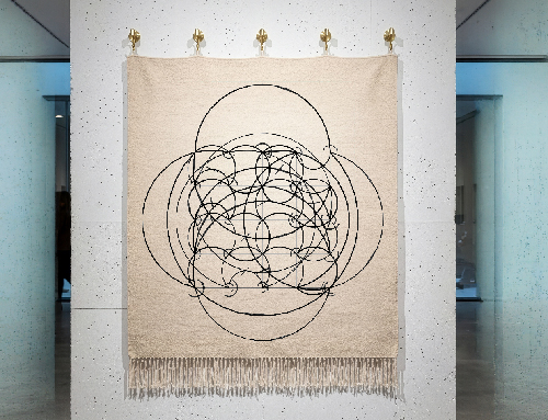 Installation view: a single magic-square arc motif woven as a Jacquard tapestry with a knotted fringe, hung by brass hooks on a white gallery column